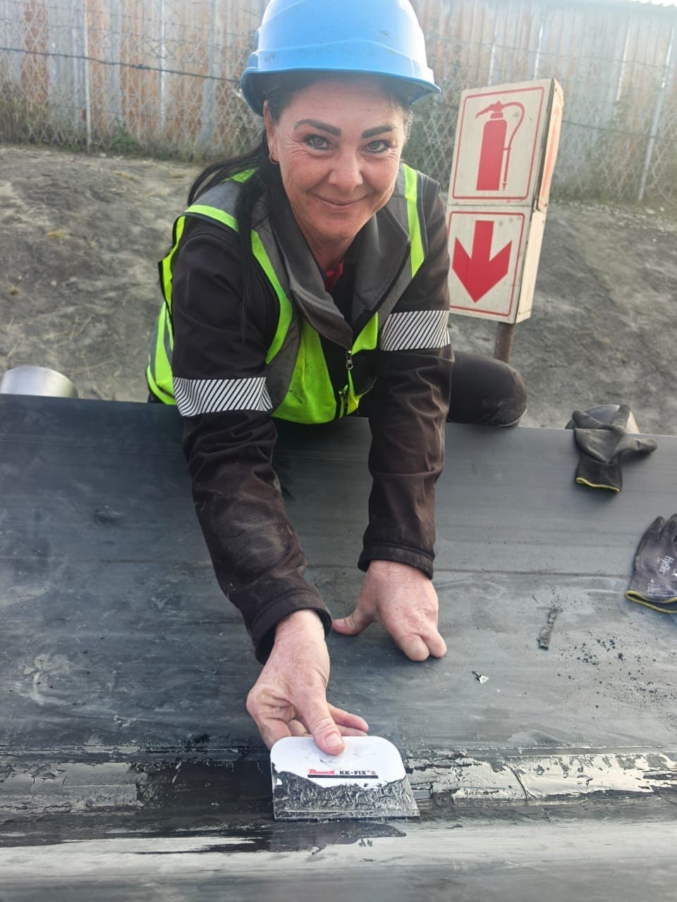 A person in a hard hat and hi-vis applying Bosworth KK-Fix compound over a belt splice with the kit spatula on a stationary belt